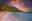 Wide sandy beach with waves gently lapping at shoreline. Beyond are palmtrees and forested mountains under a fiery red sky dotted with clouds. People are swimming just out from shore and colourful painted boats are moored beyond